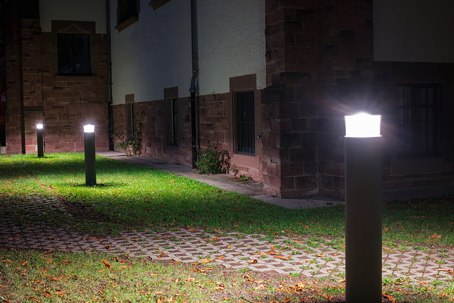 Beside a red brick campus building, three bollards light a dusty pathway