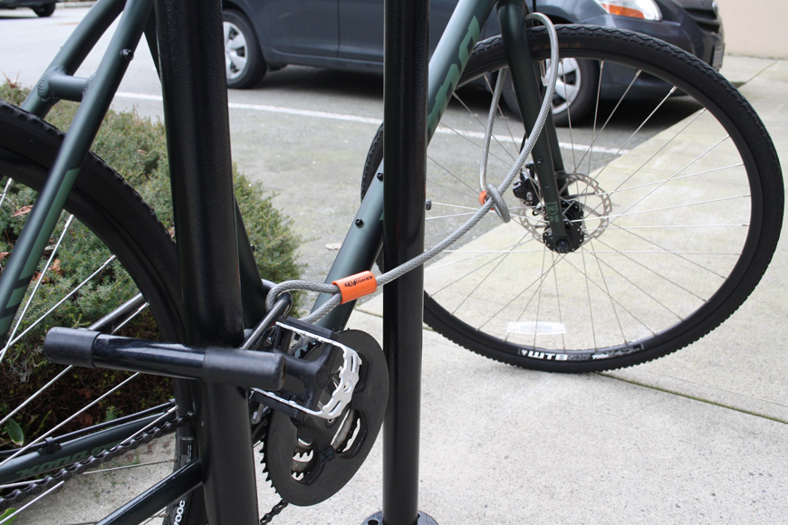 A dark green bike is locked with a u-lock in the back triangle, cabled to the front wheel