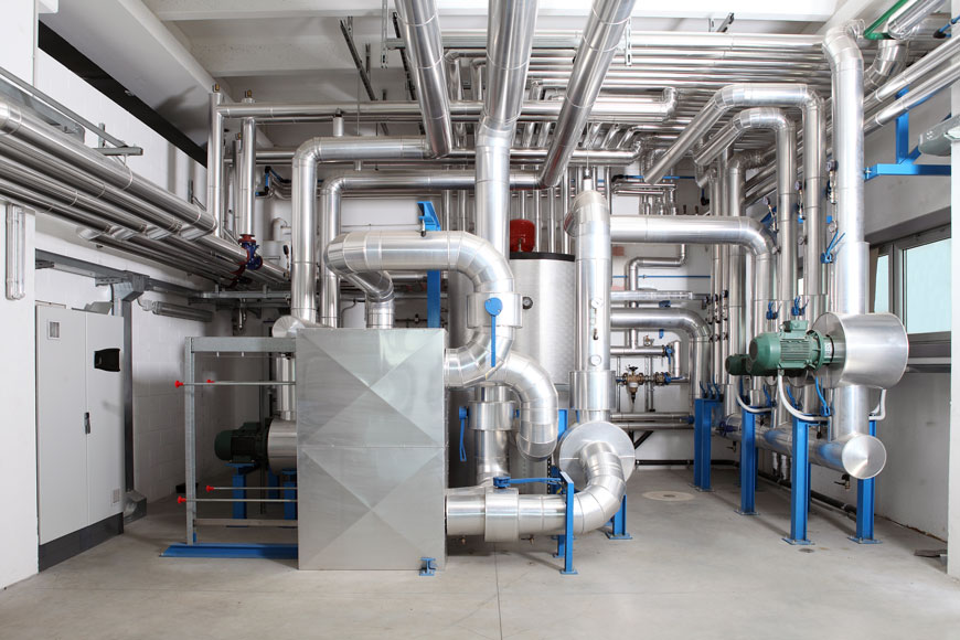 A shining series of pipes leads from a boiler; blue bollard-rails protect people and the piping.